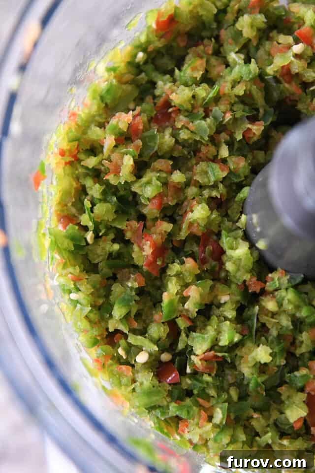 A food processor bowl filled with a colorful mix of finely chopped red and green bell peppers and jalapeños.