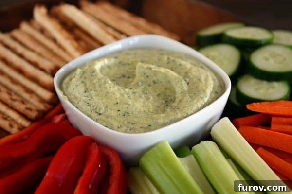 A vibrant bowl of zucchini hummus surrounded by a colorful array of fresh cut vegetables like carrots, bell peppers, cucumbers, and crispy pretzel sticks, perfect for dipping.