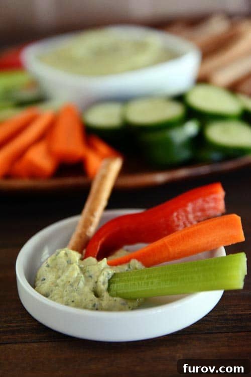 A close-up shot of a small white ramekin filled with creamy zucchini hummus, garnished with a drizzle of olive oil, accompanied by fresh carrot and celery sticks.