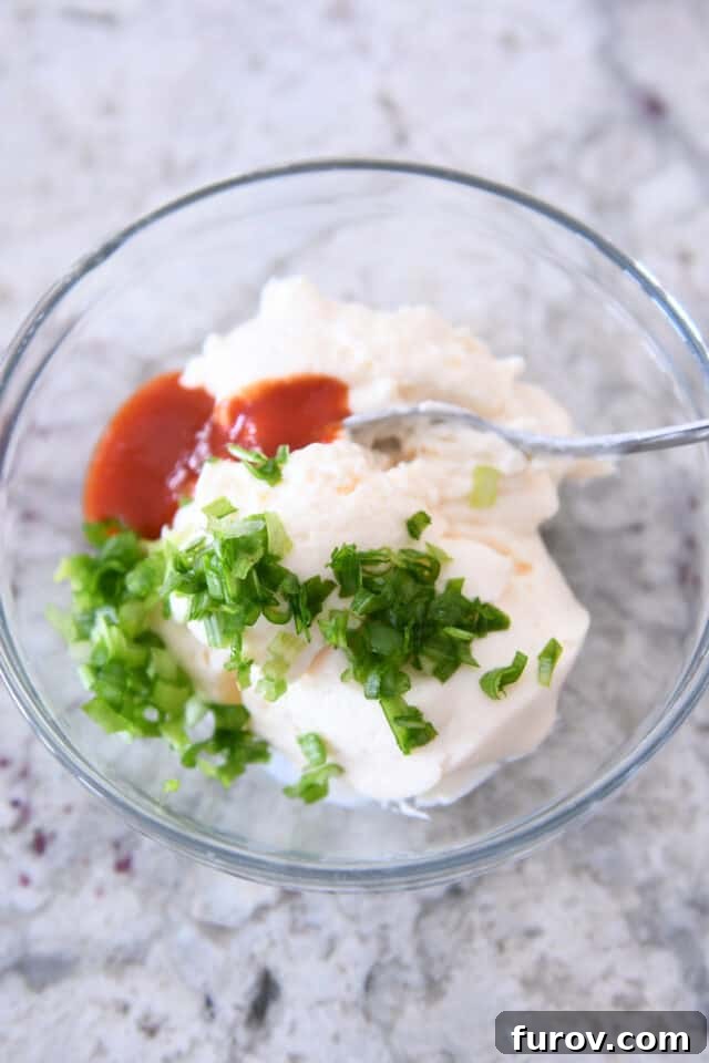 Glass bowl containing creamy sauce ingredients: mayonnaise, sour cream, finely chopped green onions, and a swirl of sriracha.