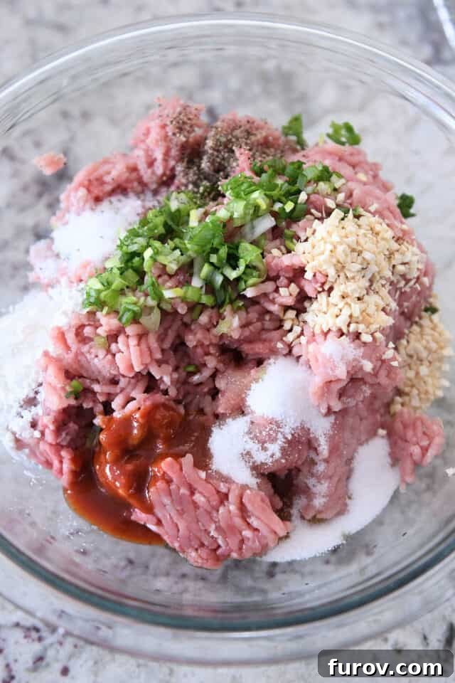 Glass bowl filled with ingredients for meatballs: ground turkey, ground pork, green onions, minced garlic, ginger, and sriracha.