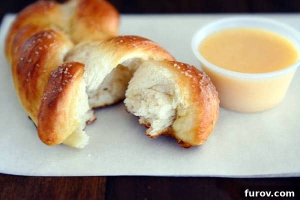 A perfectly braided pretzel twist with a piece broken off, resting beside a bowl of vibrant, smooth cheese sauce.