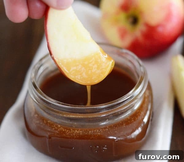 A crisp apple slice being dipped into a jar of thick, homemade caramel sauce.