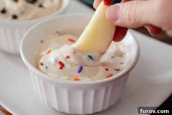 A crisp apple slice being gently dipped into a small white ramekin filled with a colorful funfetti dip, adorned with sprinkles.