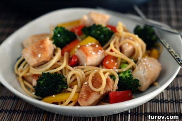 A white bowl of cooked chicken and vegetable lo mein topped generously with toasted sesame seeds.