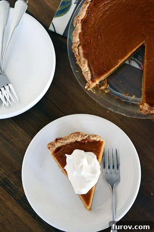 A perfectly baked classic pumpkin pie with a slice removed, revealing its creamy texture, served on a white plate with a dollop of whipped cream.