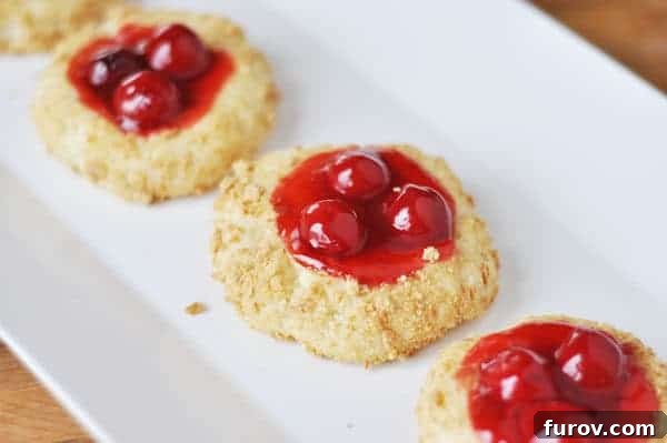 Cherry cheesecake cookies lined up neatly on a white plate, showcasing their inviting appearance with golden crumbs and red cherry centers.