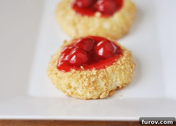 A close-up view of a single cherry cheesecake cookie, showing its crumb-coated exterior and a vibrant cherry filling nestled in the center.