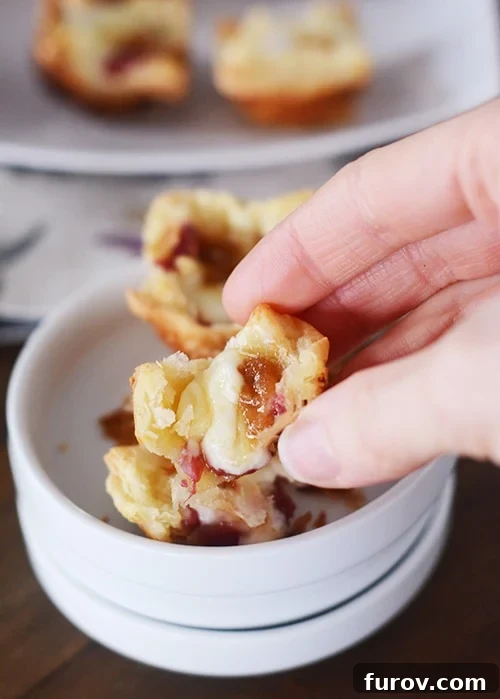 A puff pastry brie bite broken in half, revealing gooey cheese and jam, with other bites in a bowl below it, showcasing their irresistible texture and filling.
