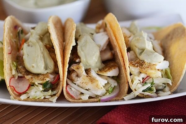 Flavorful and Fast Broiled Tilapia Tacos Ready in 30 Minutes 3 Three corn tortilla broiled tilapia tacos with fresh toppings, including a vibrant slaw and avocado sauce, lined up side-by-side on a white platter, ready to be served.