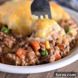 A generous serving of Shepherd's Pie in a white bowl, with a fork digging in to reveal the perfect layers of savory beef and cheesy mashed potatoes.