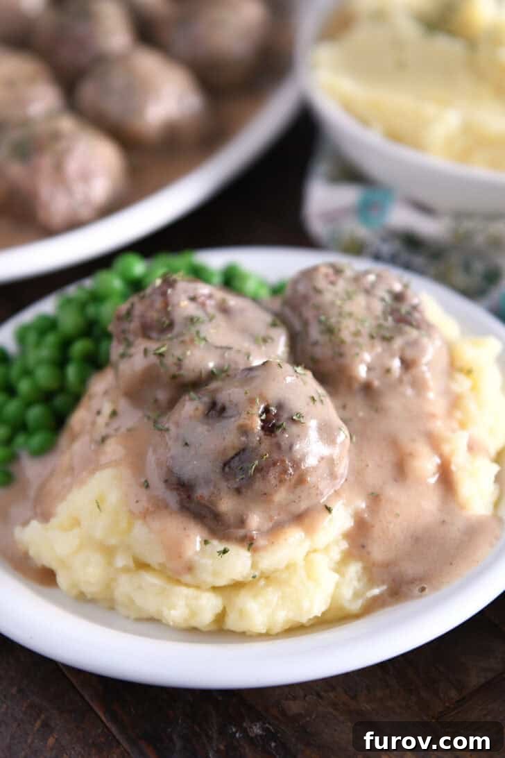 Creamy Skillet Swedish Meatballs 2 Three golden brown Swedish meatballs artfully placed on a mound of creamy mashed potatoes, generously smothered in a luscious, thick brown gravy, garnished with fresh parsley.