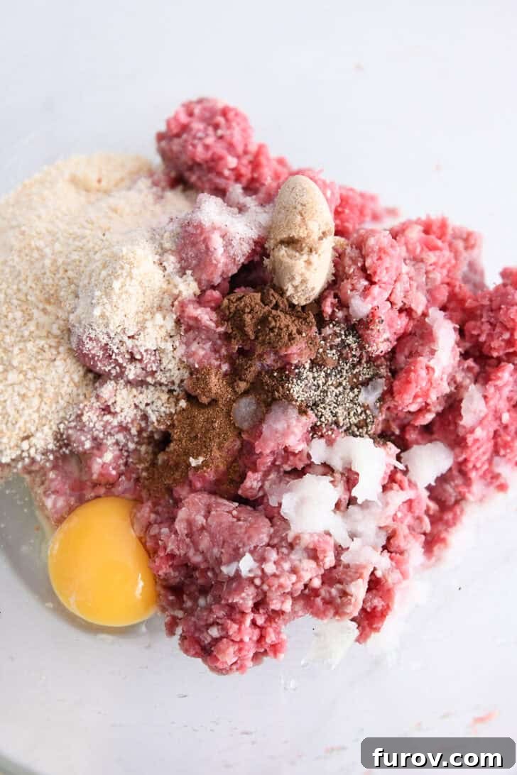 Creamy Skillet Swedish Meatballs 3 Glass bowl filled with the prepared meatball mixture, showcasing ground beef, grated onion, a whisked egg, pepper, nutmeg, brown sugar, and bread crumbs, ready to be formed into meatballs.