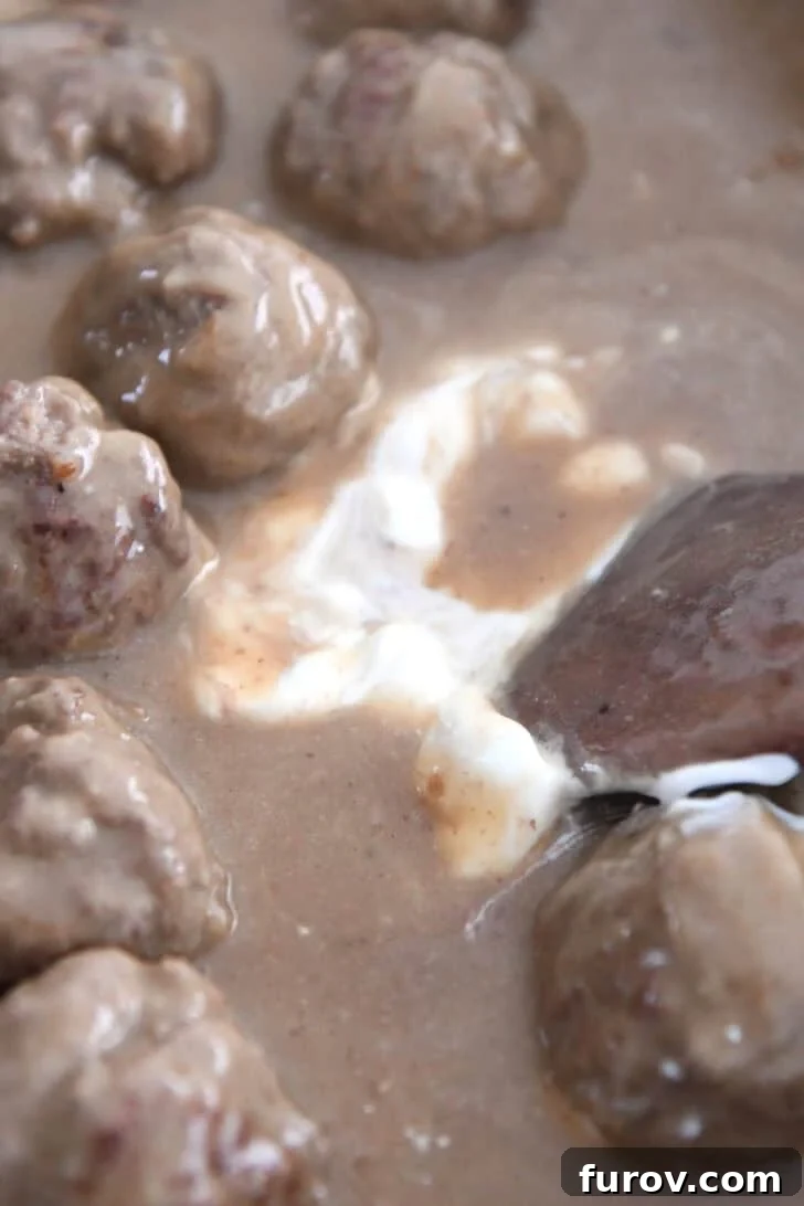 Creamy Skillet Swedish Meatballs 8 A hand gently swirling sour cream into the simmering brown gravy for Swedish meatballs in an electric skillet, creating a creamy, tangy finish.