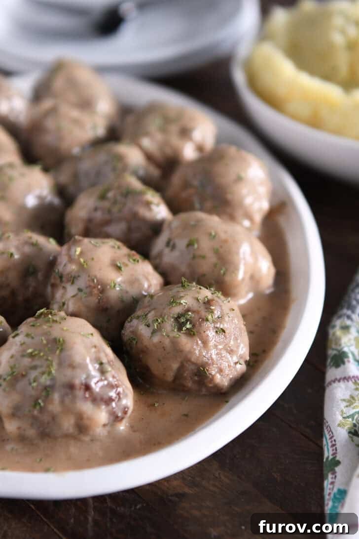 Creamy Skillet Swedish Meatballs 9 A pristine white platter generously piled with golden brown, cooked Swedish meatballs, lavishly covered in a thick, glossy brown gravy, and elegantly garnished with finely chopped dried parsley.