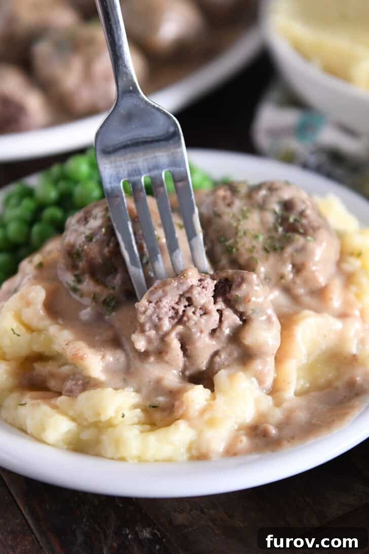 Creamy Skillet Swedish Meatballs 10 A fork piercing a succulent, half-cut Swedish meatball, resting on a bed of creamy mashed potatoes, all richly coated in the signature brown gravy. A perfect bite.