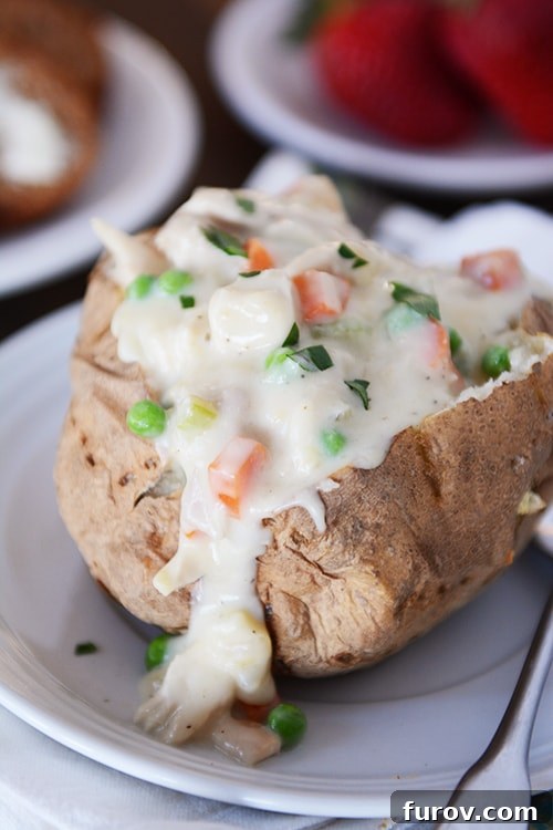 A baked potato on a white plate split open and filled with creamy chicken pot pie sauce, vibrant vegetables, and tender chicken pieces.