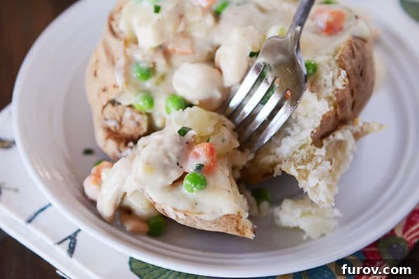 A baked potato generously topped with a creamy chicken pot pie sauce, featuring visible pieces of chicken and vibrant vegetables, with a fork poised to take a bite.