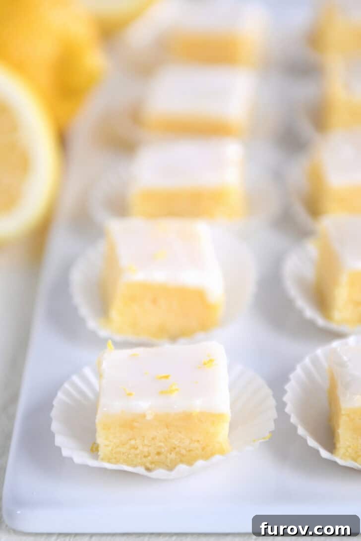 A line of lemon brownie squares topped with lemon zest and a white glaze in white muffin cups on a white serving platter, ready to be enjoyed.