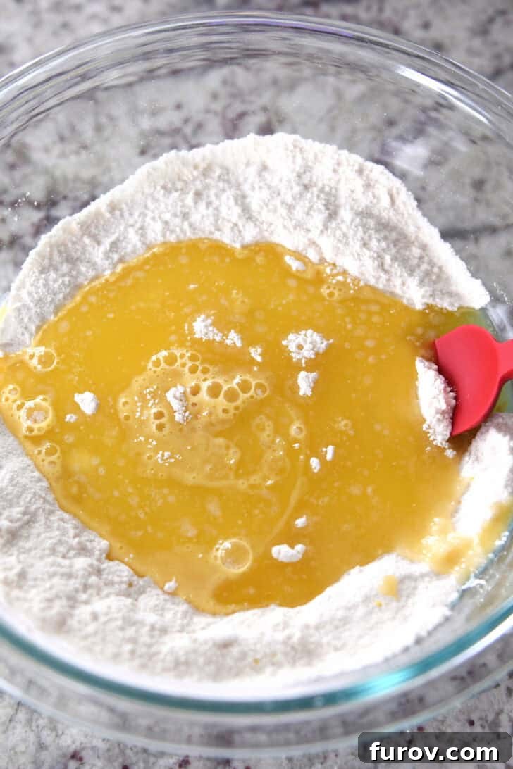 Top down view of a glass bowl with flour, a lemon and egg mixture in the middle of the flour, and a red spatula on the side, showcasing the initial mixing stage of the lemon brownie batter.