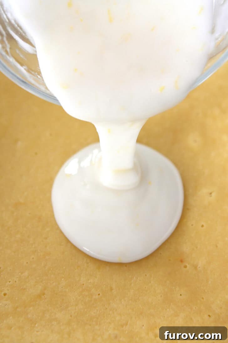 A glass bowl pouring a thick white glaze over a pan of freshly baked lemon bars, illustrating the glazing process.