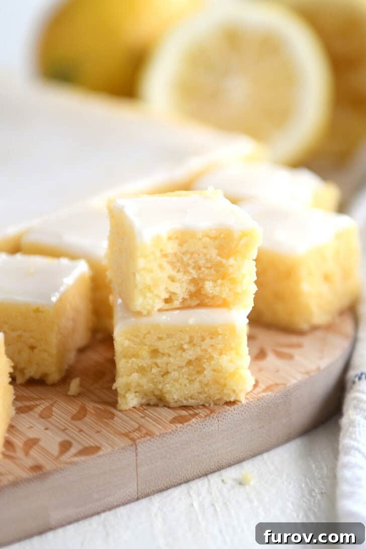 Two small, perfectly glazed lemon brownie squares, stacked invitingly on a rustic wooden cutting board, with more lemon brownie squares blurred in the background, hinting at abundance.