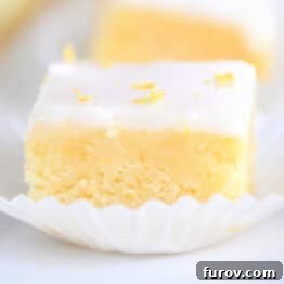 A single lemon brownie square, topped with a delicate white glaze and a sprinkle of lemon zest, resting in a pristine white muffin liner, showcasing its perfect individual serving size.