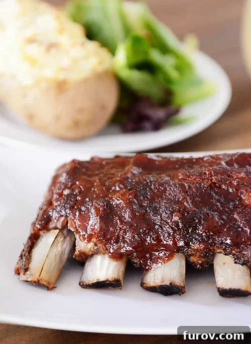 Slow Cooker Fall-Off-The-Bone Root Beer BBQ Ribs 2 A rack of tender baby back ribs, generously smothered in a rich, glistening Root Beer BBQ sauce, presented beautifully on a white platter. A side of roasted potatoes and a fresh green salad complete the meal.