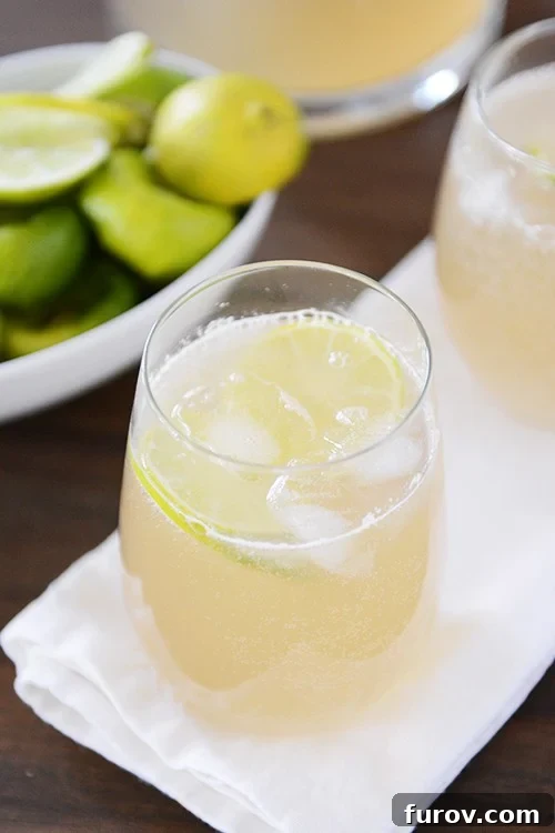 Top view of two glasses of iced limeade in front of a white bowl of sliced limes, garnished with fresh mint.