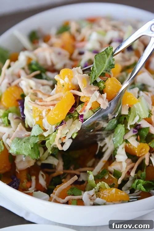 Tongs taking a generous serving of mandarin orange chicken salad from a large white bowl, highlighting the fresh, colorful ingredients.