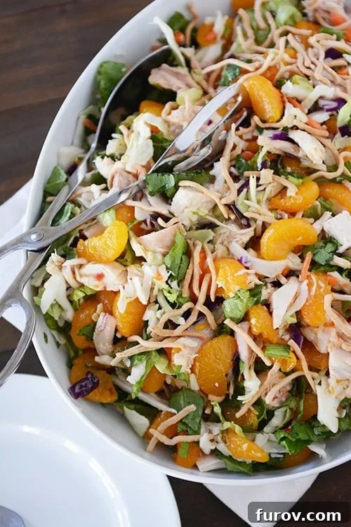 A large white platter displaying a bountiful Mandarin Orange Chicken Salad with a pair of serving tongs, garnished with fresh green onions.