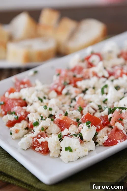 A white tray of Greek feta dip with sliced baguettes in the background, garnished with fresh herbs and a drizzle of olive oil.