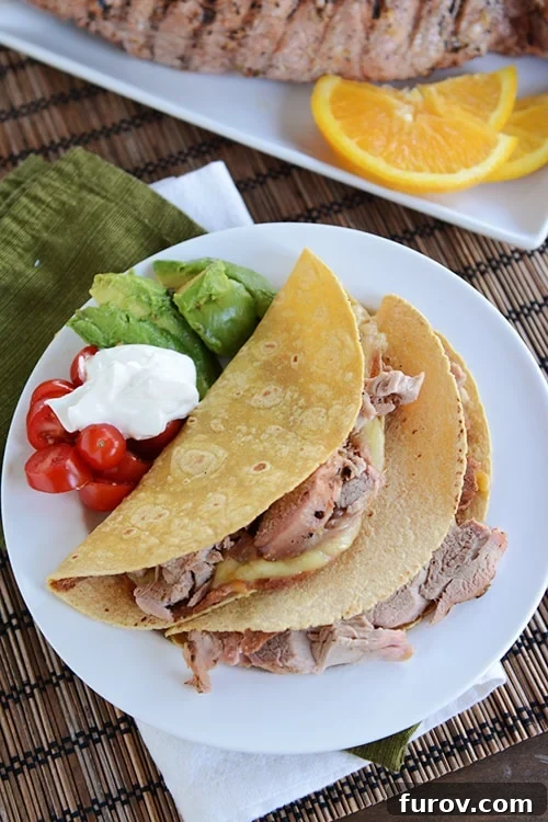 Two grilled mojo pork tacos served on a white plate, garnished with fresh cherry tomatoes, a dollop of sour cream, and diced avocado.
