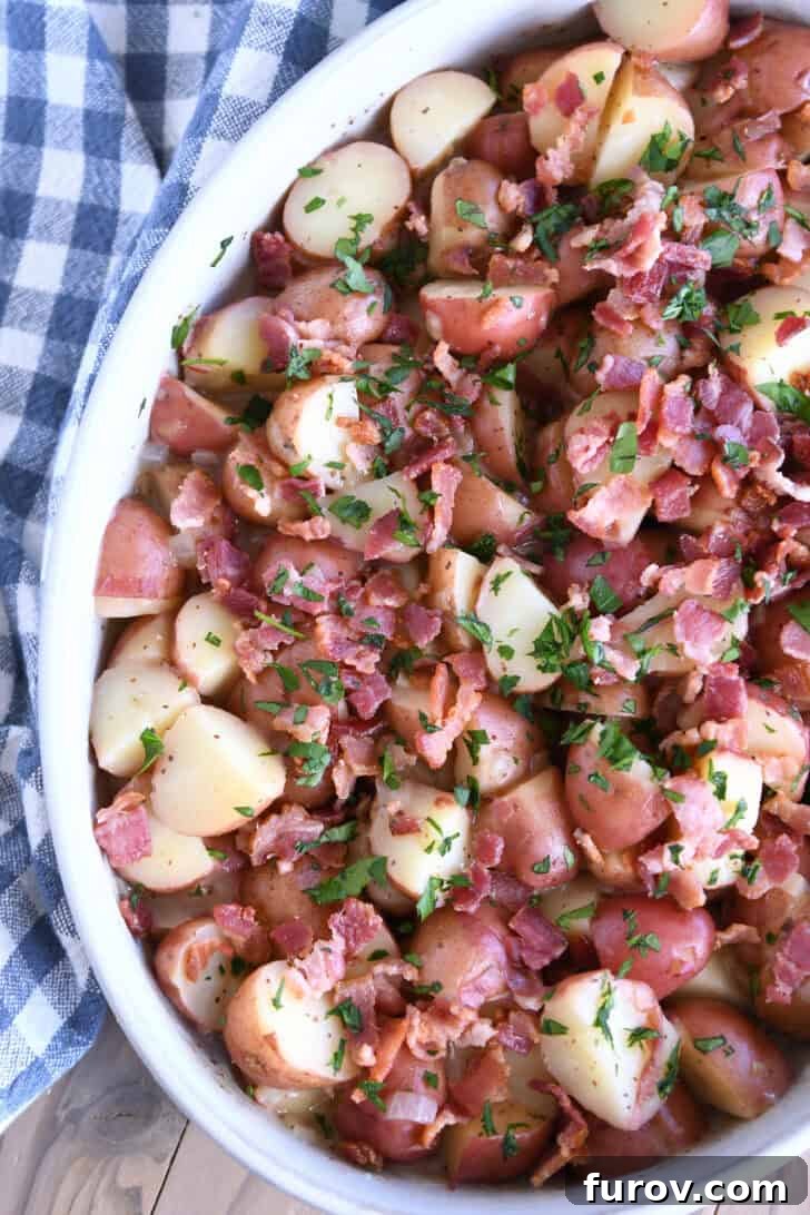 Top down view of a white oval dish with potato chunks, bacon pieces, and chopped parsley, showcasing a vibrant German potato salad.