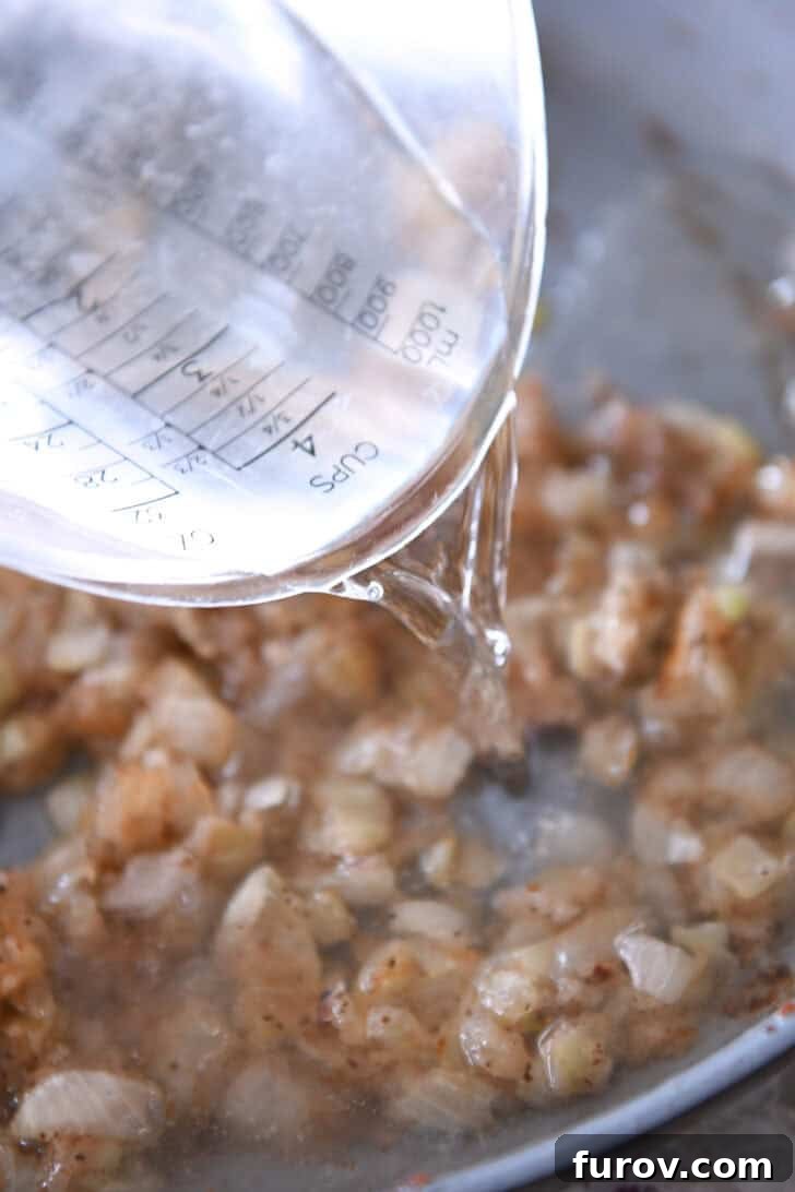 A liquid measuring cup pouring water into a skillet with browned onion pieces and the roux, showing the start of liquid addition.
