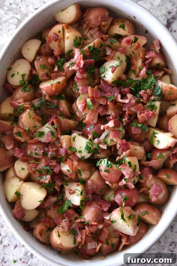 Top down view of a white oval dish with potato chunks, bacon pieces, and chopped parsley, ready for serving.