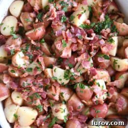 Top down view of a white oval dish with potato chunks, bacon pieces, and chopped parsley, representing the German Potato Salad recipe.