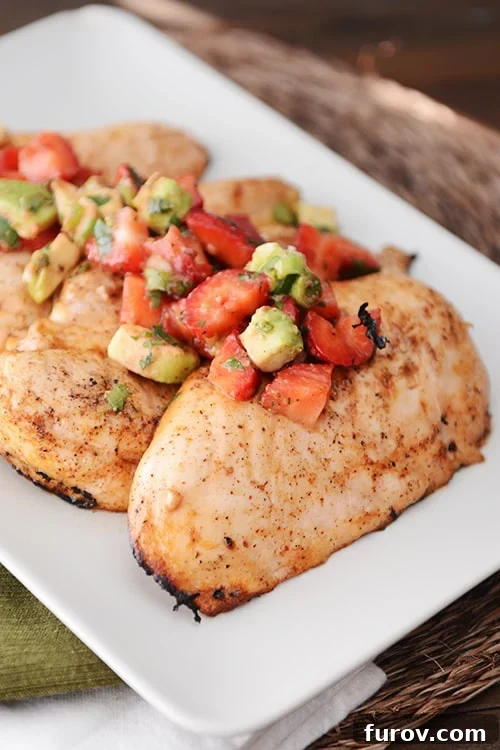 A white tray of perfectly grilled chicken breasts topped generously with a fresh, colorful strawberry-avocado salsa.
