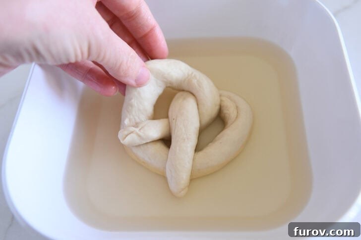 Pillowy Soft Homemade Pretzels 5 An unbaked, formed pretzel dough being dipped into a shallow water bath on a baking sheet.
