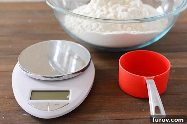 A kitchen scale, red measuring cup, and clear glass bowl full of white flour. 