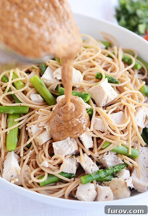 Thai Peanut Sesame Chicken Noodles 2 A bowl of cooked pasta, chicken cubes, and asparagus pieces getting sauce poured over the top.