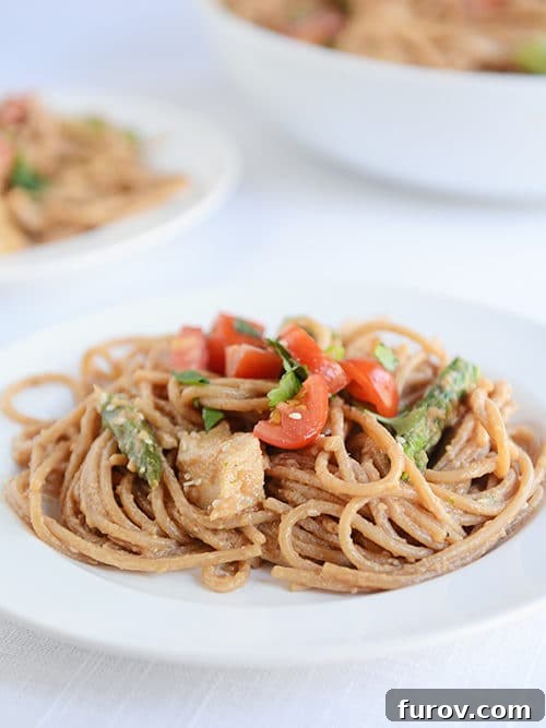 Thai Peanut Sesame Chicken Noodles 4 A white plate of cooked spaghetti noodles topped with asparagus and tomato pieces.