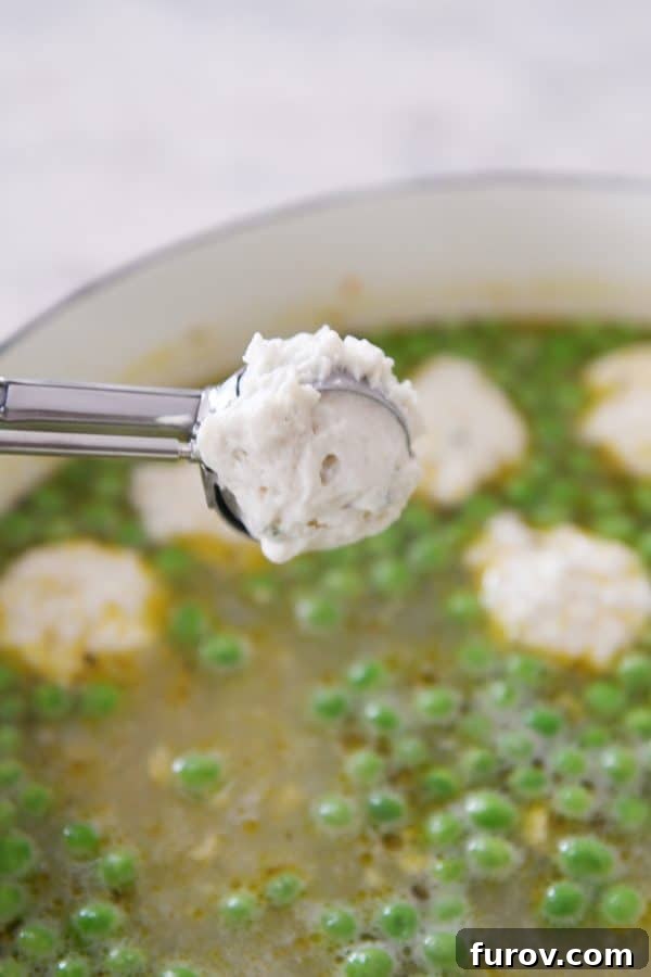 Cozy Chicken and Veggie Dumpling Soup 4 A hand using a small cookie scoop to drop dumpling batter into a pot of simmering soup.