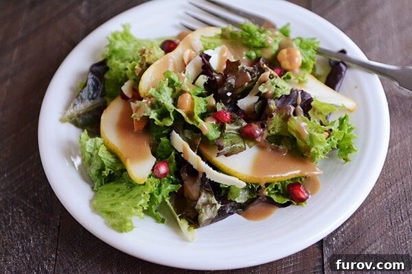A white plate with green salad topped with sliced pears, pomegranates, and dressing, elegantly arranged.