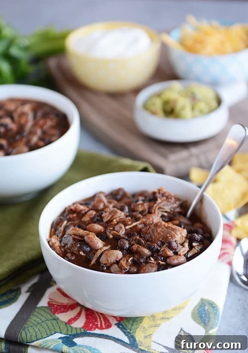 A steaming white bowl of pressure cooker chicken and black bean chili, garnished with fresh cilantro, a dollop of sour cream, and a spoon. Bowls of vibrant toppings like shredded cheese and avocado are softly blurred in the background.