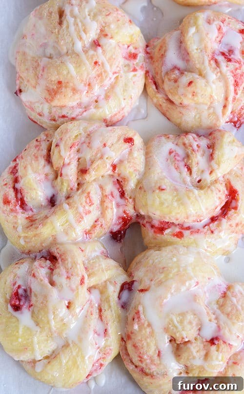 Six, baked strawberry cream cheese rolls on a piece of parchment paper drizzled with a white glaze.