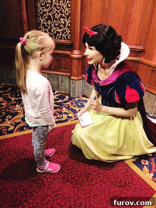 A little girl smiling next to a Disney princess character, getting an autograph.