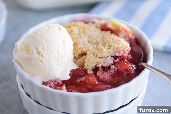 A white ceramic ramekin holds a portion of strawberry cobbler with a perfect golden biscuit topping and a scoop of melting vanilla ice cream on the side, ready to be enjoyed.