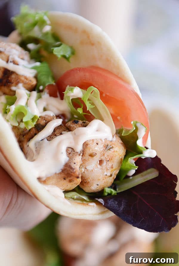 A hand holding a flatbread wrap filled with grilled lemon chicken, crisp lettuce, juicy tomato slices, and drizzled generously with spicy garlic sauce.