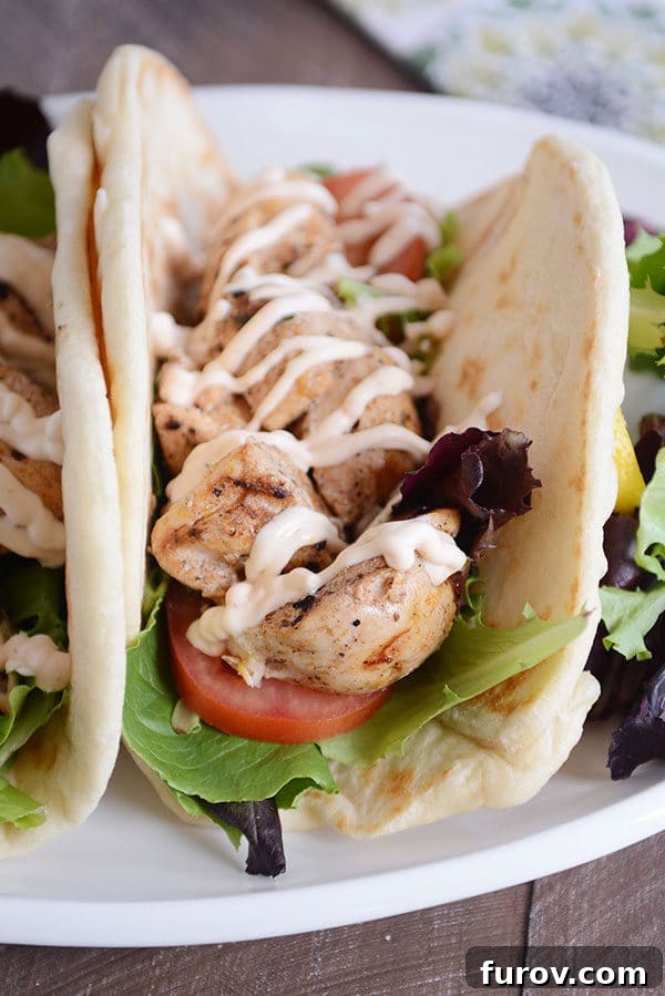 A close-up shot of a single grilled lemon chicken wrap, showcasing the tender chicken, fresh tomato, crisp lettuce, and generous drizzle of creamy spicy garlic sauce.